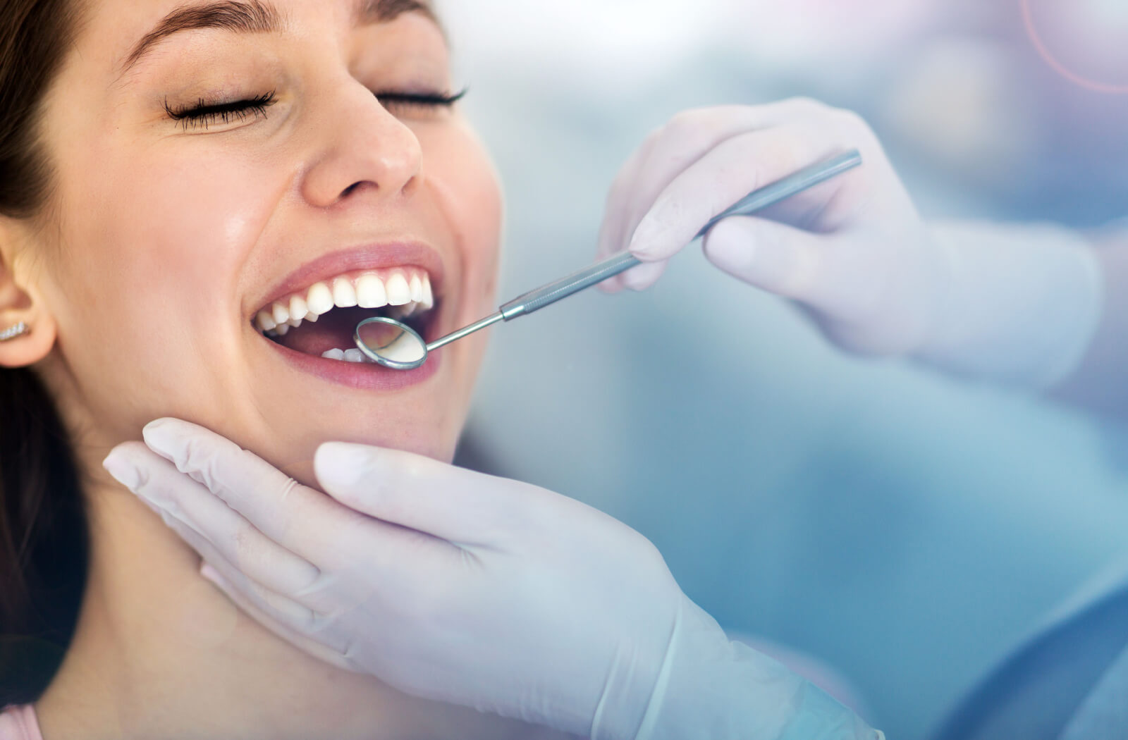 Close-up of a young woman smiling as a dentist examines her teeth with a dental mirror
