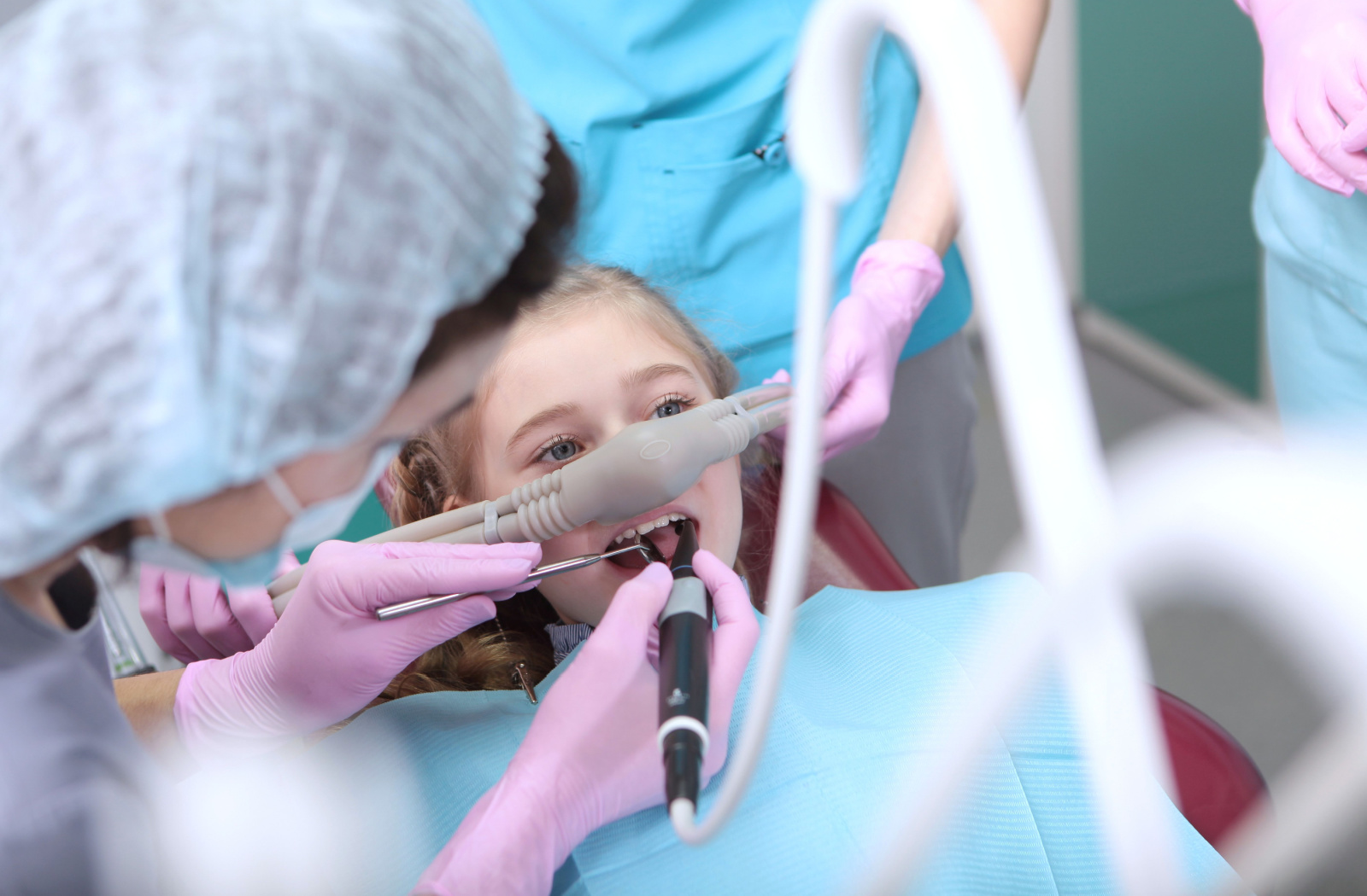 A female child is in a sedated state, wearing a sedation mask, as she undergoes a dental cleaning administered by a woman dentist.
