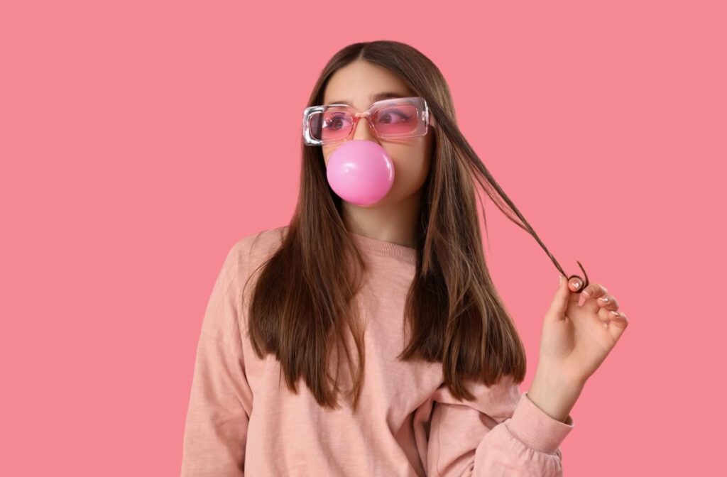 Against a pink background, a teenager twirls a strand of hair while blowing a large pink bubble of bubblegum.