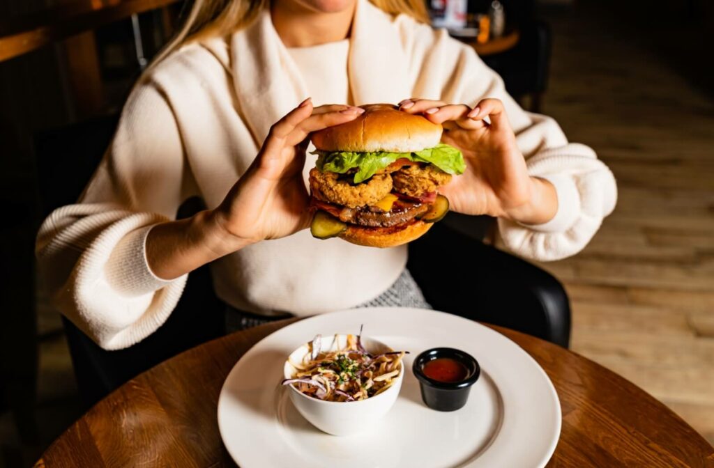 A woman at a restaurant holding a large gourmet burger, illustrating the freedom to eat different foods by removing Invisalign aligners before meals.
