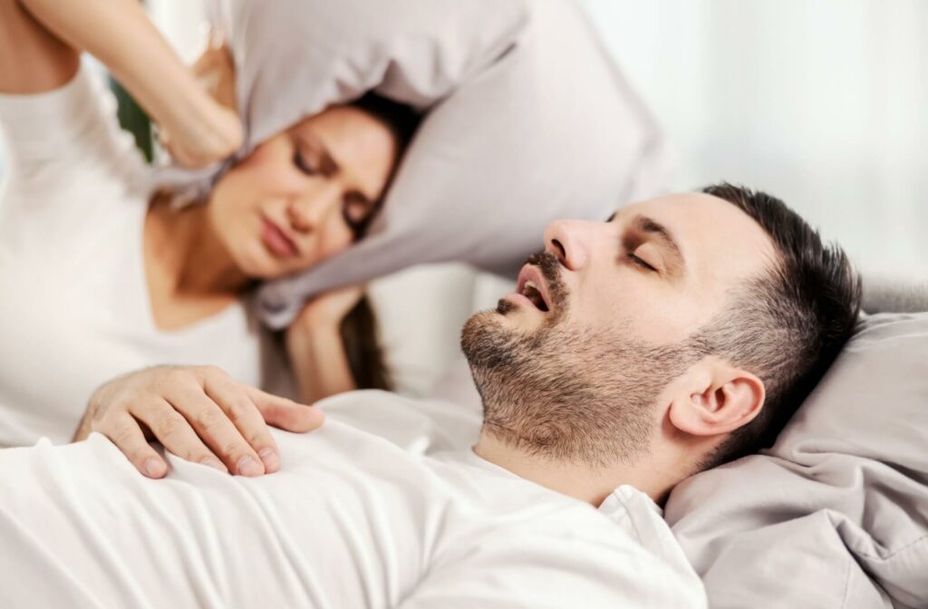 An adult pulling a pillow around her ears in frustration as her partner snores due to his sleep apnea.