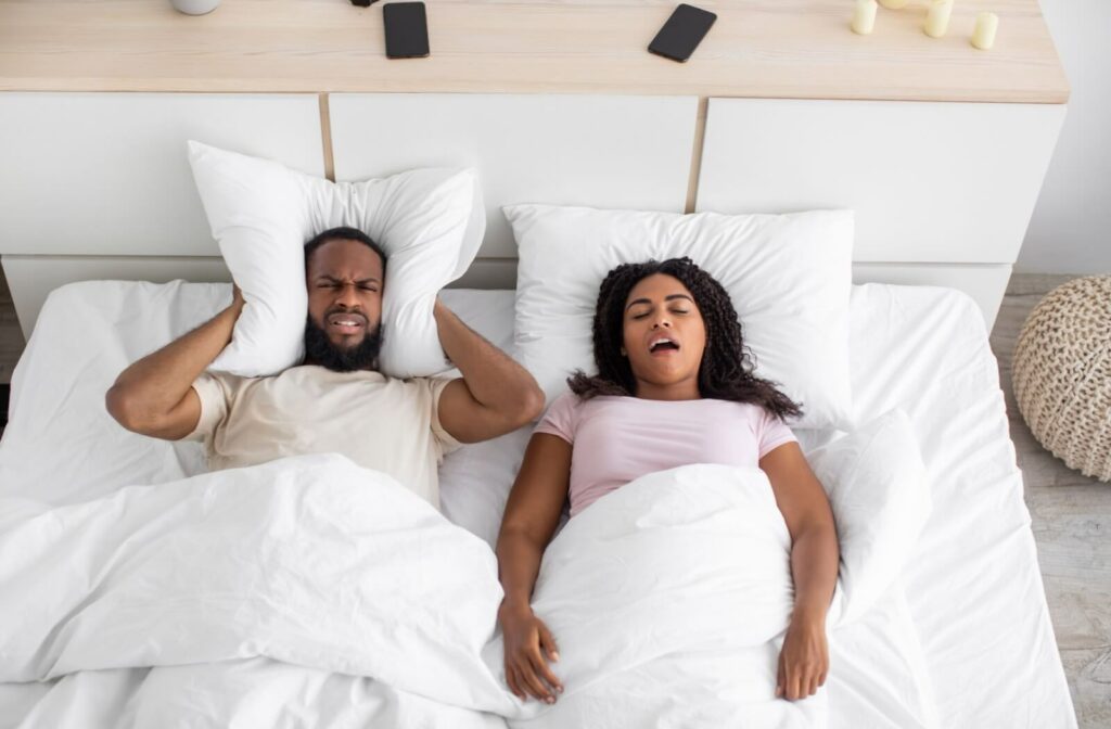Man covering ears while woman snores in bed due to sleep apnea.