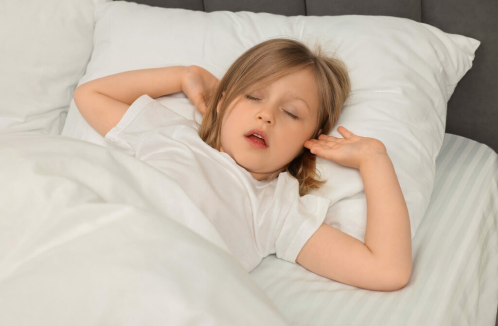 A young girl sleeps with her mouth open and arms raised beside her head, possibly indicating restless or labored sleep.