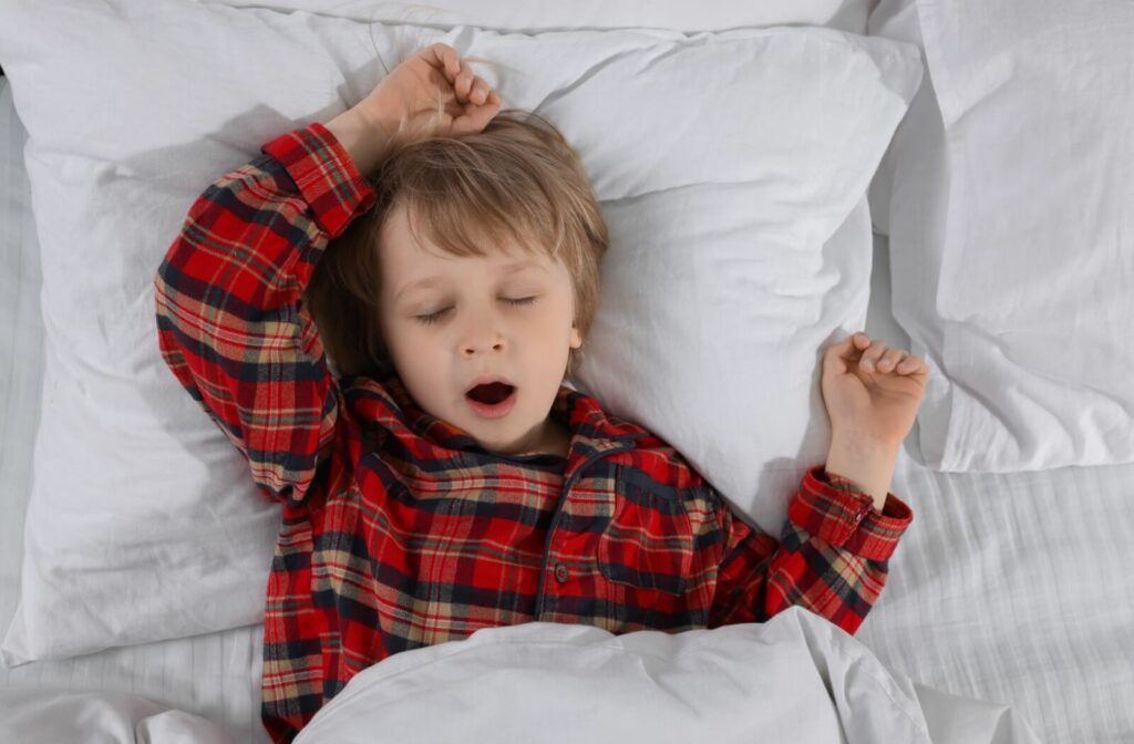 Young child in red plaid pajamas yawning while lying in bed with white pillows.