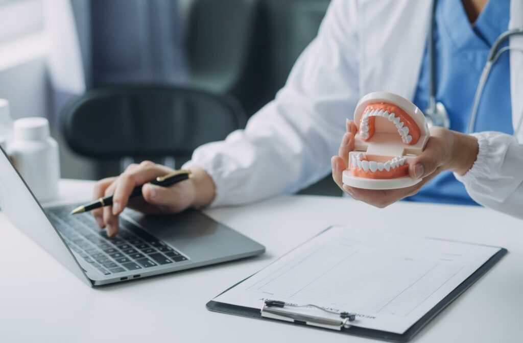 Dentist holding a dental model of teeth in one hand while using a laptop with the other, with a clipboard and pen on the desk.