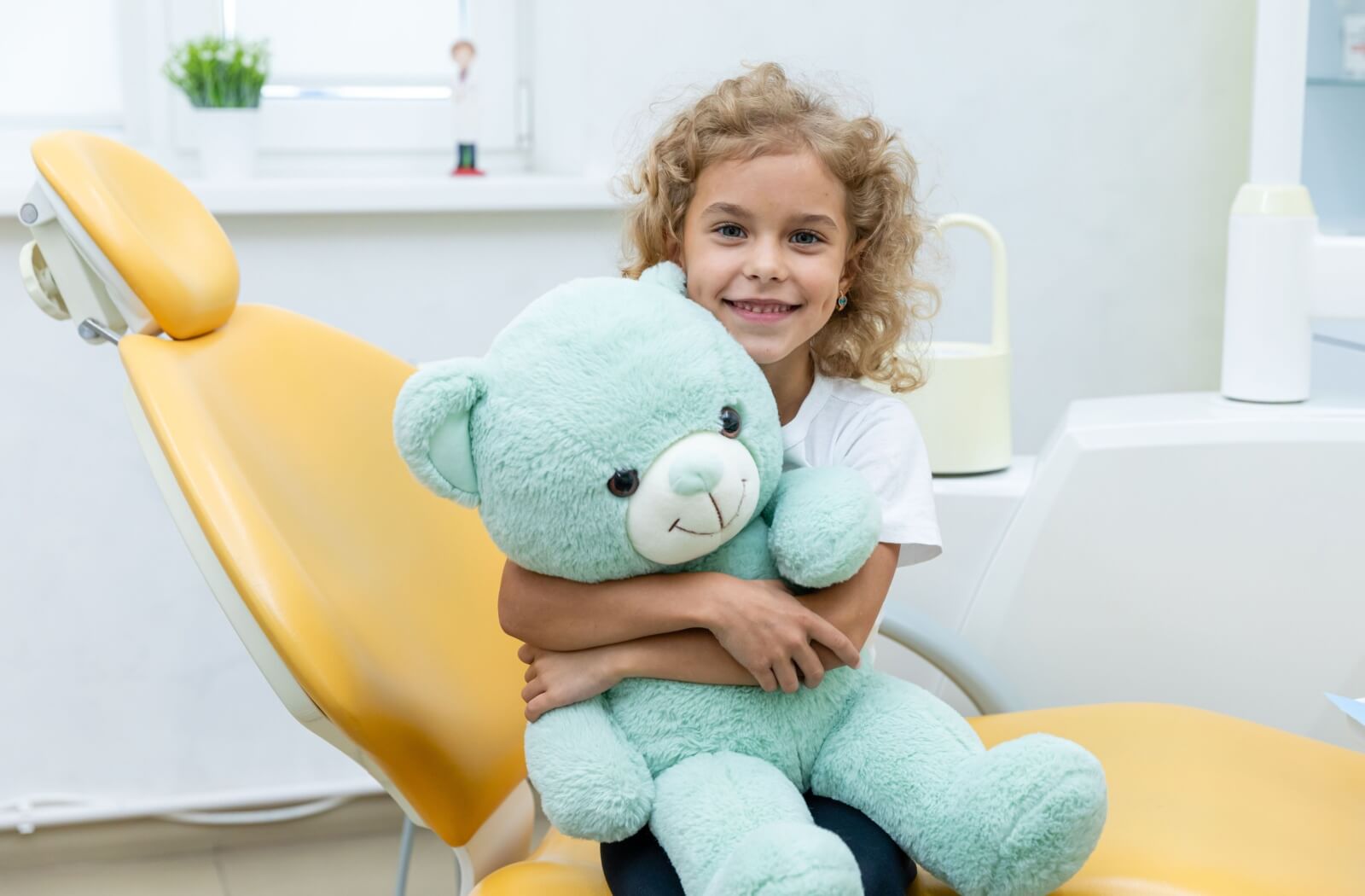 Smiling young child sitting in a dental chair, hugging a large light blue teddy bear in a brightly lit dental office.