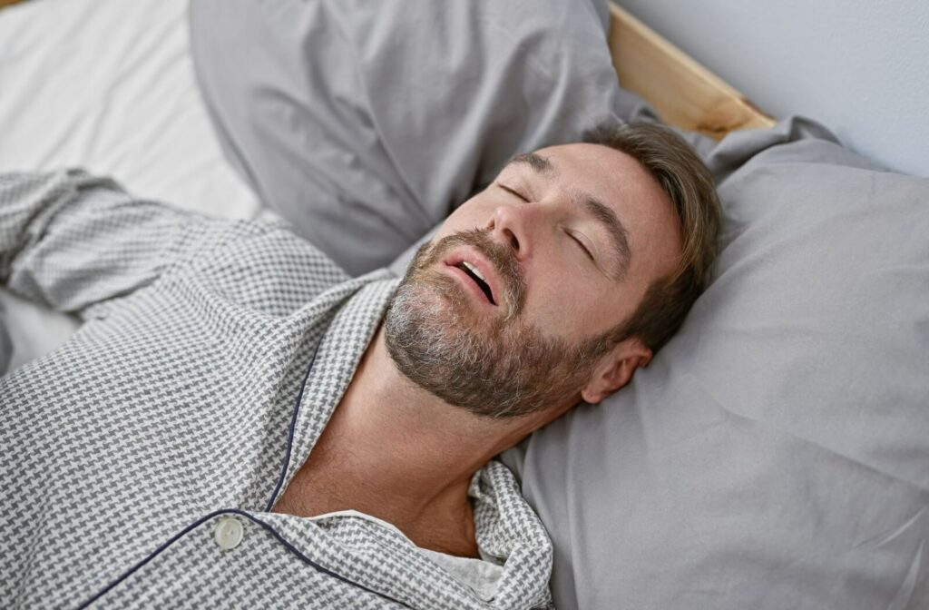 Man sleeping on his back with his mouth open, resting on grey pillows and wearing patterned pajamas.