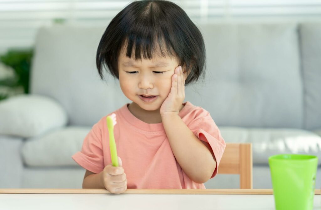 A young child holding a toothbrush and touching their cheek with a pained expression, illustrating a toothache from decay.