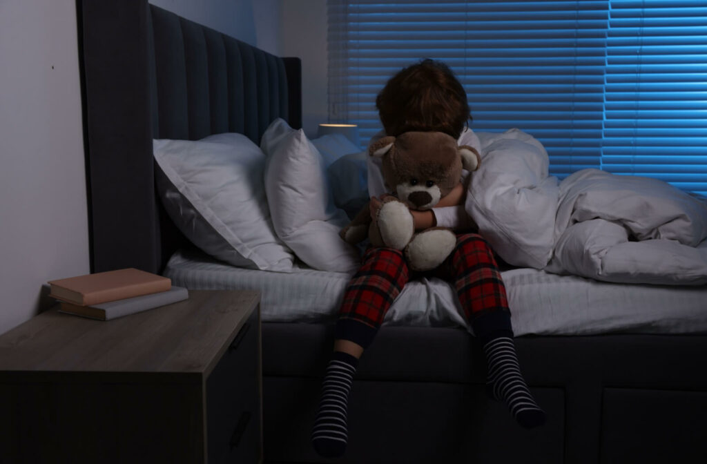 A child sitting up in bed at night holding a teddy bear.