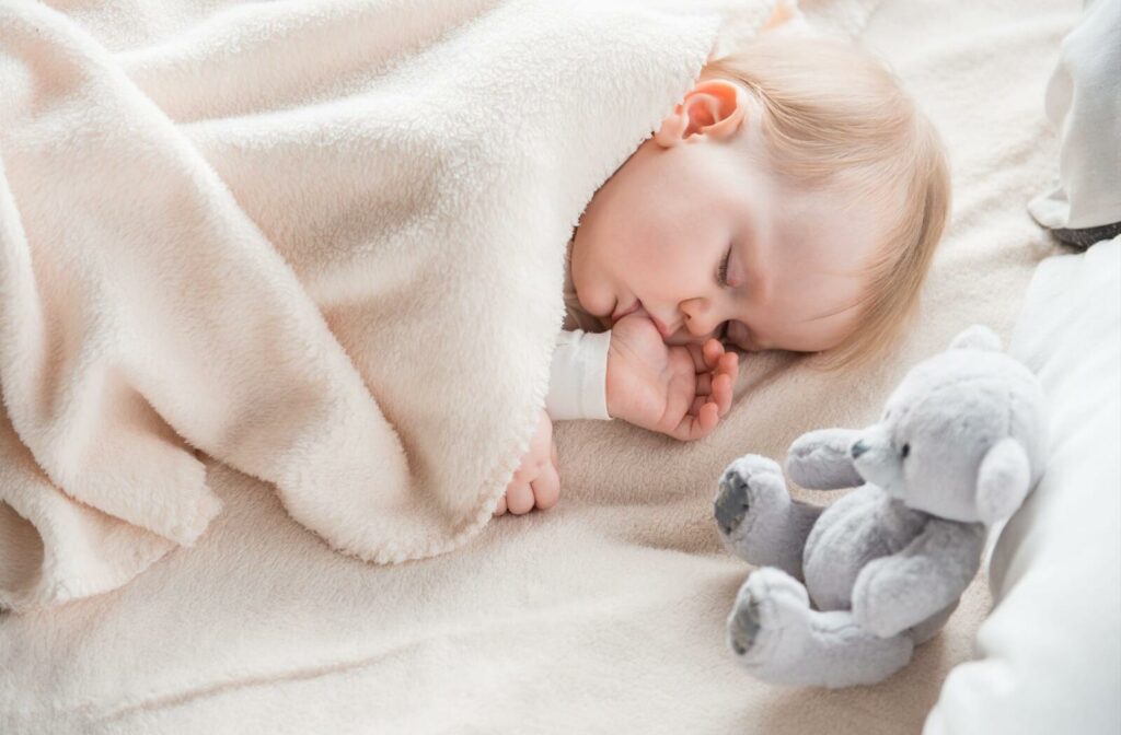 A close-up of a sleeping toddler sucking their thumb while resting under a soft cream blanket.